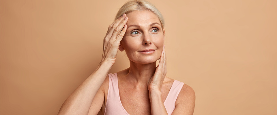 A imagem mostra o rosto e parte do tronco de uma mulher de pele branca e olhos claros, que aparenta ter cerca de cinquenta anos. Seus cabelos são loiros e estão presos para trás. Com uma das mãos, ela toca a testa, enquanto com a outra, toca uma das bochechas. Ela olha para o lado direito da tela, enquanto esboça um leve sorriso, sem mostrar os dentes. Veste uma regata rosa-claro. O fundo da imagem é rosa pastel.