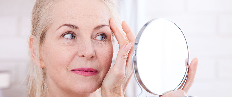 A imagem mostra o rosto de uma mulher branca de olhos claros e cabelos loiros. Ela aparenta ter cerca de cinquenta anos, e seus cabelos estão presos. Ela segura um espelho redondo em uma das mãos, e com a outra, toca uma de suas sobrancelhas, enquanto encara o espelho. O fundo da imagem é claro, e está desfocado.